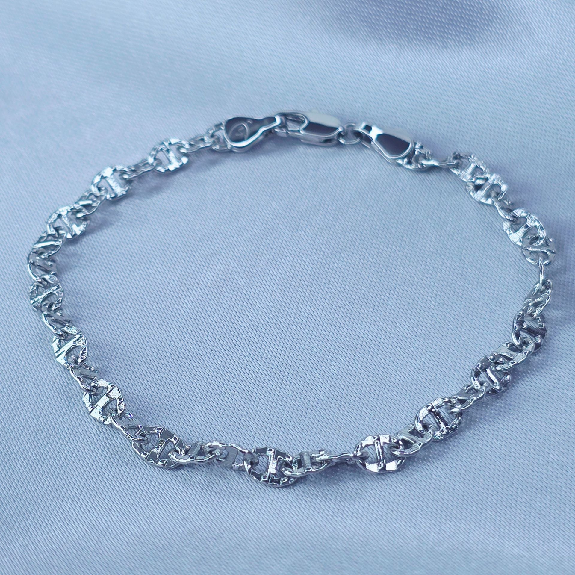 A close-up view of a White Gold Plated  anchor mariner link bracelet with textured links on a soft, reflective, light-blue fabric surface – Aurora Jewelry.