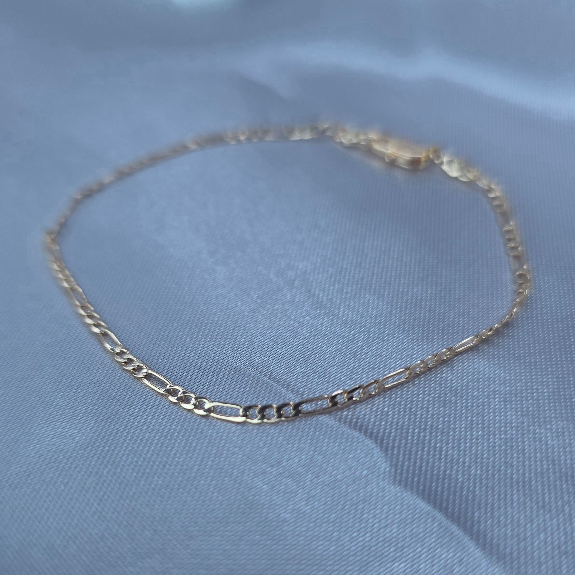High-shine gold-plated Figaro link bracelet displayed on a white satin fabric background – Aurora Jewelry Collection